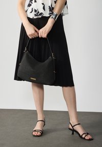 Svart vävd hobo-väska med en texturerad yta, guldaccenter, bärs tillsammans med en veckad svart kjol och sandaler med remmar i svart.