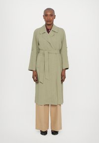Cappotto lungo verde chiaro con cintura in vita, maniche ampie e colletto. Abbinato a pantaloni beige a gamba larga e scarpe in tessuto nero.