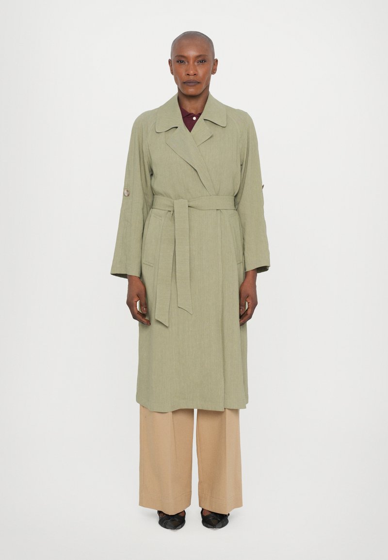 Cappotto lungo verde chiaro con cintura in vita, maniche ampie e colletto. Abbinato a pantaloni beige a gamba larga e scarpe in tessuto nero.
