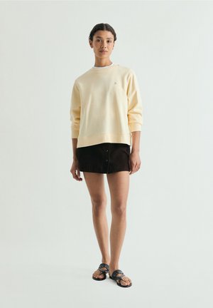 Frau trägt hellgelben Sweatshirt, schwarzen Minirock mit Knöpfen und schwarze mit Nieten verzierte Sandalen, steht vor einem schlichten hellen Hintergrund.