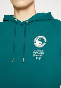 Tyrkysová mikina s klokaním vreckom, šnúrkami so zlatými koncami a kruhovým bielym logom s textom a symbolmi na hrudi.