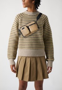 Borsa a spalla beige e nera con due tasche con zip; indossata sopra un maglione a righe e una gonna plissettata color kaki. Tessuto morbido e design elegante.