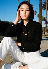 Fiatal nő rövid fekete hajjal keresztbe tett lábakkal ül a járdán, fekete kabátot, fehér nadrágot és fehér tornacipőt visel, háttérben pálmafák.