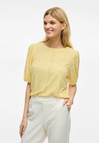 Blouse jaune à manches courtes avec un motif de perforation en pois et un col rond. Les manches sont bouffantes, associées à un pantalon de couleur claire.