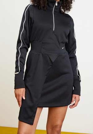 Personne portant une robe noire à manches longues avec un passepoil blanc sur les manches, une fermeture éclair à l'avant et un ourlet asymétrique dévoilant une jambe.
