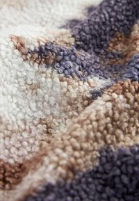 Gros plan d’un tissu doux à texture bouclée avec un mélange de couleurs beige, blanc et violet dans un motif abstrait.