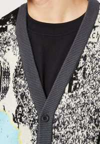 Cardigan en tricot avec un motif texturé monochrome, col côtelé gris et fermeture à un bouton, superposé sur une chemise noire.