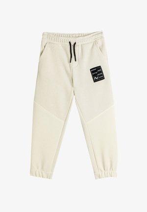 Pantalones de chándal color crema con tejido texturizado, cinturilla elástica, cordón negro y un parche cuadrado negro en el muslo izquierdo.
