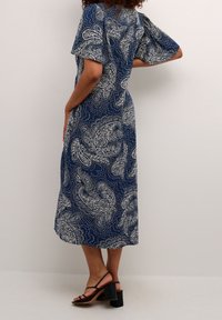 Žena v midi šatech námořnické modré barvy s bílými paisley vzory, stojící s jednou rukou na boku, v černých sandálech na blokovém podpatku.