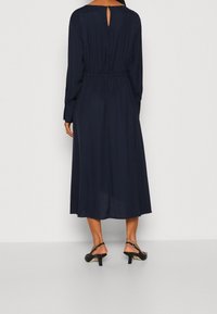 Žena v modrých midi šatech s dlouhým rukávem a malým klíčovým otvorem na zádech, obutá v černých slingback kočičích podpatcích, stojící zády.