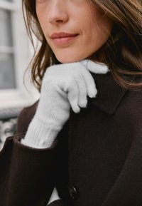 Guanti bianchi lavorati a maglia con polsini a coste appoggiati su un cappotto marrone scuro, che mostrano una texture morbida e una vestibilità aderente intorno al polso.