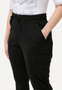 Pantaloni cargo neri con vita elastica, chiusura con cordoncino, tasche laterali e un tessuto testurizzato. Presenta una tasca cargo funzionale.
