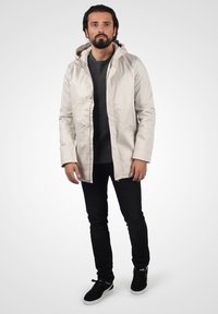 Lysfarget, vanntett jakke med hette, trykknapper og lange ermer, kombinert med svarte jeans og svarte sneakers.