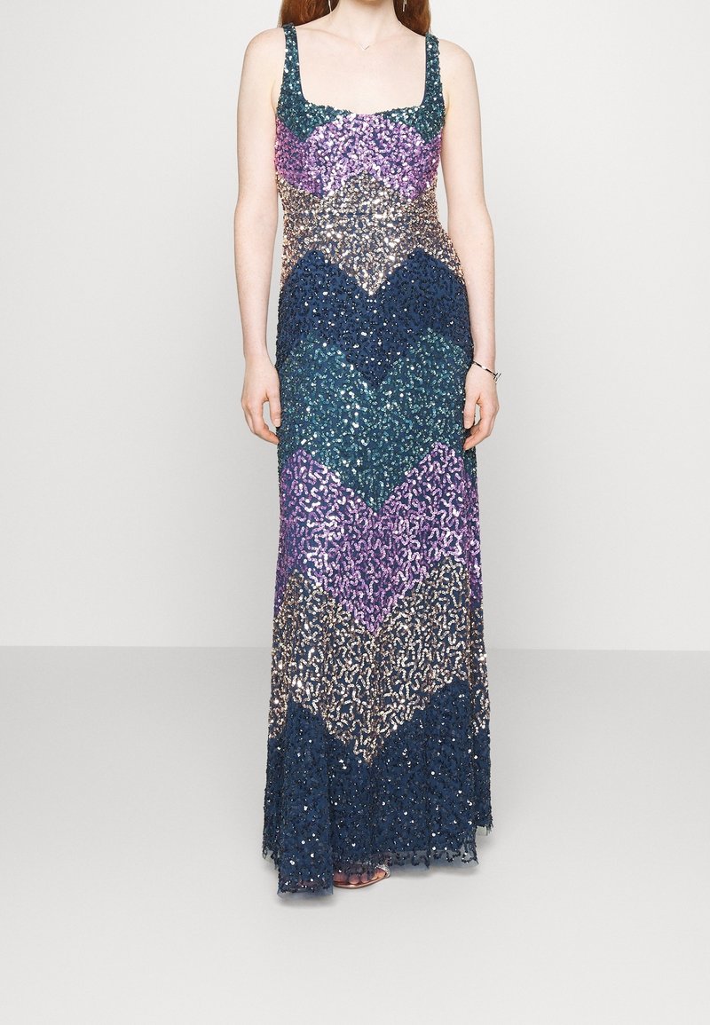 Femme portant une robe longue sans manches ornée de sequins avec des motifs en chevrons dans des tons violet, bleu et or sur un fond uni.