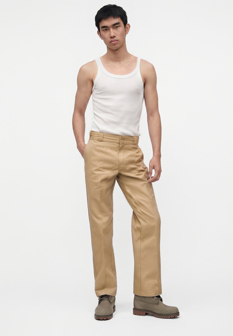 Giovane uomo in piedi con le mani in tasca, indossa una canottiera bianca, pantaloni beige e stivali grigi su uno sfondo bianco semplice.