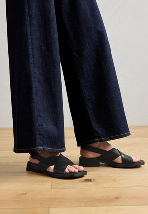 Sorte lædersandaler med krydsede remme, flad sål og åben tå, sat sammen med vidde bukser i mørk denim på et trægulv.