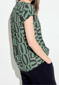 Top de manga corta en verde con un estampado abstracto negro que presenta grandes cifras. Escote redondeado, corte relajado y tejido texturizado.