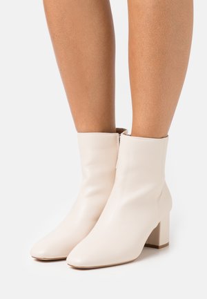 Beżowe botki sięgające do kostki, wykonane z gładkiej skóry, z kwadratowym noskiem i masywnym obcasem, posiadające średniej wysokości cholewkę oraz minimalne przeszycia.