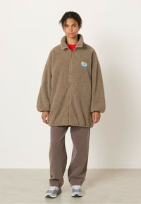 Fleece-takki vaaleanruskeana, joissa on vetoketju edessä ja suuri kaulus. Takissa on myös logo ja se on rento malli. Yhdistetty ruskeisiin housuihin ja harmaisiin lenkkareihin.