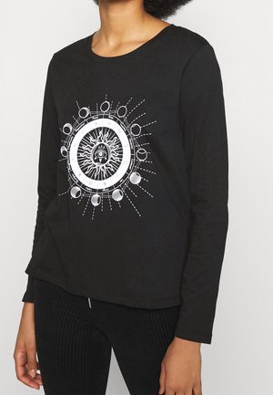 Personne portant un t-shirt noir à manches longues avec un motif circulaire blanc représentant le soleil, un œil, les phases de la lune et les symboles du zodiaque sur le devant, associé à un pantalon noir.