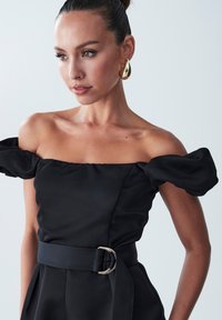 Vestido negro de un hombro con mangas abullonadas y una cinturilla ancha con una hebilla circular plateada. Tejido suave y silueta estructurada.