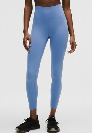 Kobieta w jasnoniebieskich legginsach z wysokim stanem i czarnych butach sportowych, stojąca na tle jednolitego białego tła.