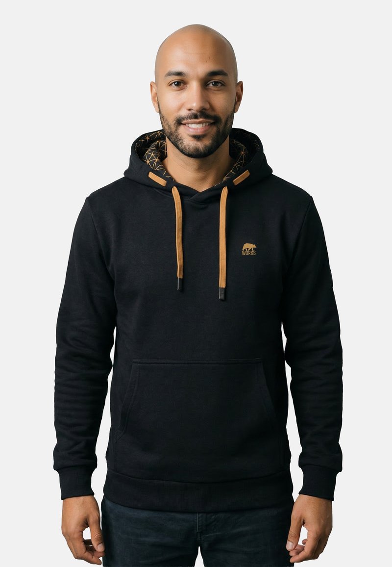 Uomo con barba che indossa una felpa nera con coulisse marroni e un piccolo logo a forma di orso, in piedi davanti a uno sfondo bianco semplice, sorridente.