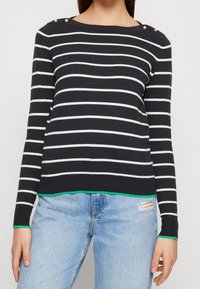 Marinrandig tröja med vita horisontella ränder, med axelknappar, grön fåll och tighta ärmar, kombinerad med ljusblå jeans.