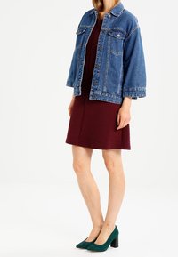 Jeansjacka med klassisk skärning, blå färg och knappstängning, i kombination med en vinröd ribbad klänning och gröna högklackade skor.