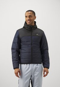 Padded jacket z czarną górną częścią i ciemnoniebieską dolną częścią. Posiada kaptur, zamek błyskawiczny oraz małe zielone logo na klatce piersiowej.