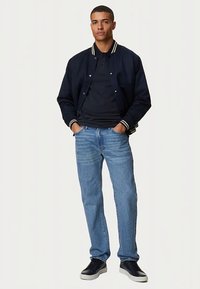 Marineblaue Bomberjacke mit weißen gestreiften Bündchen, schwarzes Poloshirt, hellblaue gerade geschnittene Jeans und schwarze Sneakers. Einfaches, lässiges Outfit.