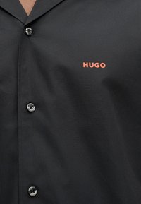 Μαύρο πουκάμισο με κουμπιά, με τρία ορατά κουμπιά και πορτοκαλί λογότυπο "HUGO" στην αριστερή πλευρά του στήθους, φορεμένο πάνω σε γυμνό δέρμα.