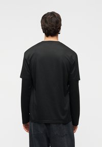 Άνδρας με καστανά μαλλιά μέτριου μήκους, ντυμένος με στρωτά μαύρα πουκάμισα και σκούρα τζιν, απεικονίζεται από πίσω μπροστά σε λευκό φόντο.