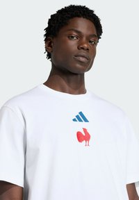 Maglietta bianca in cotone con logo Adidas blu e grafica di un gallo rosso sul davanti. Maniche corte e scollatura rotonda.