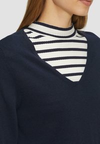Granatowy sweter z dekoltem w serek nałożony na biały sweter z granatowymi paskami na szyję. Materiały wydają się miękkie o gładkiej fakturze; prążkowany detal przy kołnierzu.