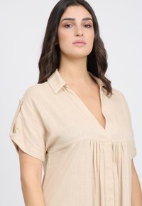 Blusa beige a maniche corte con scollo a V, fronte plissettato e vestibilità morbida. Realizzata in tessuto leggero con una sottile texture.
