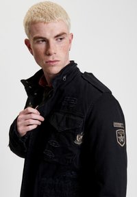 Zwarte militaire stijl jas met meerdere zakken, verouderde details en patches. Heeft een hoge kraag en rits, gemaakt van stevig materiaal.