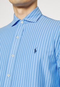 Camicia a righe azzurro chiaro con bottoni, collo classico, bottoni bianchi e un piccolo logo nero ricamato sul petto.