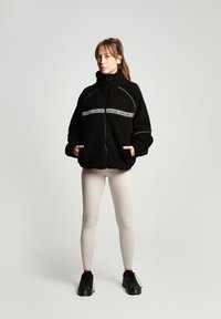 Fekete bolyhos fleece félcipzáros dzseki fehér logós csíkkal, két elülső zsebbel és magas gallérral, bézs leggings-szel és fekete sportcipővel párosítva.