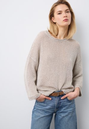 Junge Frau trägt einen beigen Strickpullover, der in blaue Jeans mit braunem Gürtel gesteckt ist, steht mit den Händen in den Taschen vor einem schlichten Hintergrund.
