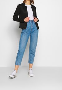 Femme en jean bleu taille haute, baskets blanches, chemise blanche et blazer noir, debout contre un mur blanc, ajustant sa veste avec les mains.
