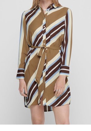 Kobieta w sukience koszulowej do kolan z długim rękawem z ukośnymi paskami w kolorach brązowym, beżowym, niebieskim i ciemnobrązowym, związanej w talii paskiem.