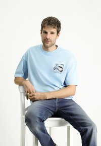 Camiseta de algodón azul claro con mangas cortas, que presenta un gráfico impreso de un scooter y el texto "Viaja Conmigo" en la parte izquierda del pecho.