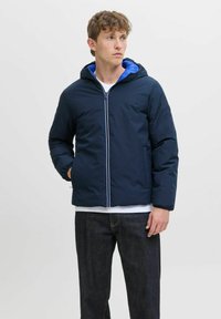 Marineblaue Steppjacke mit blauer Kapuze, vorderem Reißverschluss, Seitentaschen, elastischen Bündchen und weißem Innenfutter, kombiniert mit dunkelblauen Jeans.