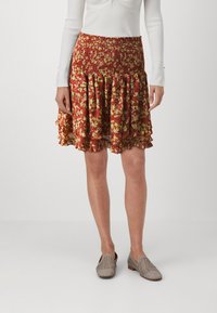 Jupe florale rouge avec des accents verts, présentant une taille smockée et un ourlet froncé. Associée à des chaussures slip-on tissées de teintes neutres.