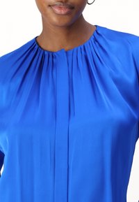 Nära bild av en person som bär en ljusblå blus med veck vid halslinjen och en dold mittsöm.