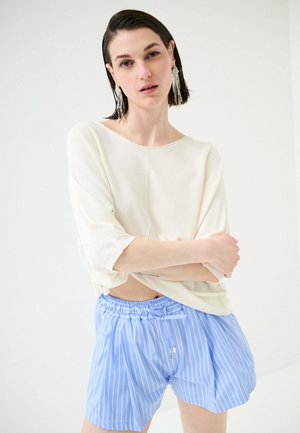 Donna con capelli corti e scuri che indossa una maglia bianca larga e pantaloncini blu a righe, in piedi con le braccia incrociate davanti a uno sfondo semplice.