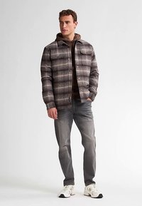 Karierte Hemdjacke in Braun- und Grautönen mit Knopfleiste vorne, zwei Brusttaschen, getragen über einem braunen Hoodie, kombiniert mit grauen Jeans und Sneakers.