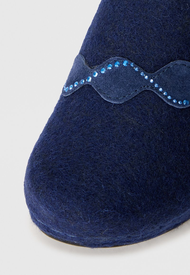Pantofole in feltro blu navale con una striscia di suede blu ondulata decorata con piccoli strass blu lungo la parte superiore. Design con punta arrotondata.