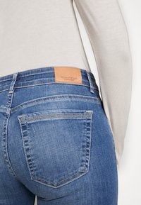 Blue jeans with a smooth denim texture, featuring a tan leather patch labeled "Marc O'Polo" on the waistband and two back pockets.